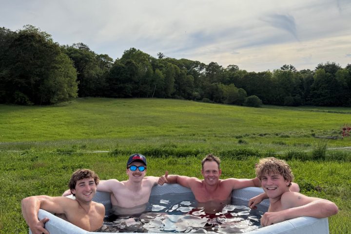 Four people smiling in an outdoor hot tub on a grassy field under a cloudy sky.